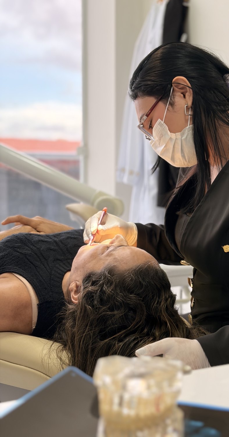 Atendimento clínico, profissional debruçada sobre a paciente, com luz natural entrando pela janela ao fundo.